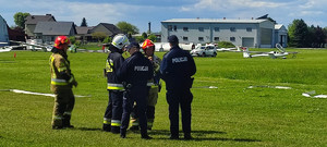 stojący na terenie lotniska obok siebie kilku strażaków i dwóch policjantów, w tle samoloty i samochód