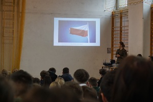 Siedzący uczniowie na hali oglądający prezentacji na której w ujęciu widac Polską flagę