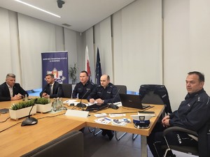 przedstawiciele Policji wraz z wójtem i przewodniczącym rady gminy siedzący przy biurku na debacie, w tle za nimi baner KPP w Krakowie