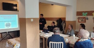 Starsze osoby siedzące przy stoliku oraz dwóch umundurowanych policjantów. Po lewej stronie ekran na którym jest wyświetlana prezentacja.