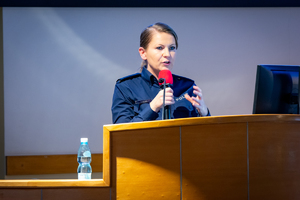 Kobieta, policjantka, prelegentka z mikrofonem w rękach stojąca przodem do zebranych.