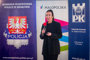 Prelegentka w ubraniu cywilnym z mikrofonem w jednej ręce na tle loga komendy wojewózkiej policji w krakowie, małopolski i politechniki krakowskiej