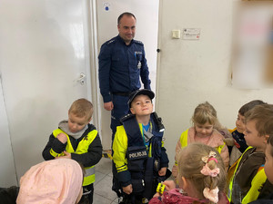 przedszkolak w policyjnej kamizelce i czapce w otoczeniu innych przedszkolaków, z tyłu stoi policjant