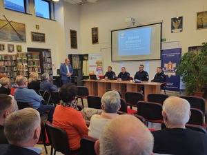 częśc uczestników debaty siedząca tyłem do zdjęcia, przed nimi stojacy wójt gminy i siedzący umundurowani policjanci, za nimi prezentacja i baner KPP Kraków