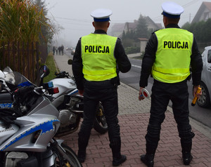 dwóch policjantów ruchu drogowego w kamizelkach odblaskowych z napisem policja stojących przy drodze tyłem do zdjęcia obok zaparkowane motocykle widoczne w częś