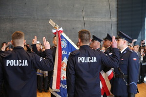 Policjanci przy sztandarze policyjnym składający ślubowanie z uniesionymi prawymi rękami i wyprostowanymi palcami wskazującym i środkowym.