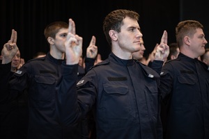 Trzech policjantów z głowami skierowanymi w lewą stronę, z uniesionymi prawymi rękami i wyprostowanymi palcami środkowym i wskazującym.