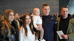 Policjant w mundurze trzymający w prawej ręce dziecko. Po jego prawej dwie kobiety, po lewej mężczyzna.