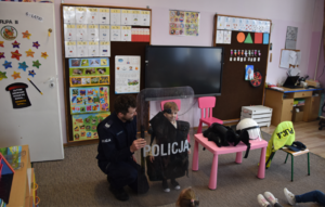 Umundurowany policjant oraz przedszkolak z elementami policyjnego umundurowania oraz tarczą z napisem Policja