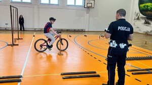 Chłopiec jadący na rowerze na sali gimnastycznej, pokonujący tor preszkód. Obok stoi tyłem umundurowany policjant ruchu drogowego, w tle kobieta.