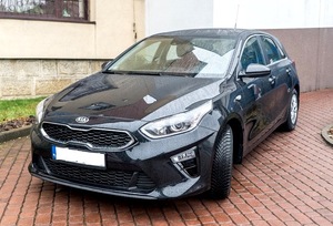 Samochód osobowy marki Kia koloru czarnego stojący przodem na kostce brukowej przed budynkiem.