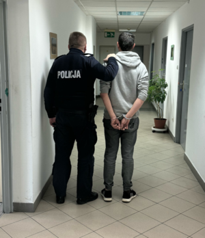 policjant stojący na korytarzu komisariatu z zatrzymanym który ma założone kajdanki na ręce trzymane z tyłu, widoczni od tyłu (2)
