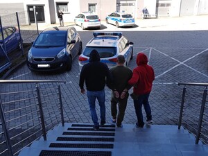 osoba zatrzymana z założonymi kajdankami na rece trzymane z tyłu  jest sprowadzana ze schodów przez funkcjonariuszy Policji w umundurowaniu cywilnym