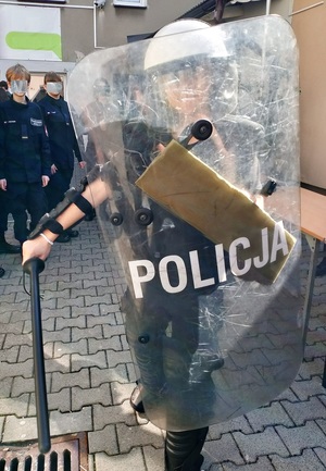 Na zdjęciu osoba ubrana w elementy stroju policyjnego, tryzmajaca w reku tarcze z napisem Policja.