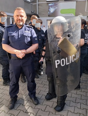 Po lewej stronie umundurowany policjant , po prawej osoba ubrana w elementy policyjnego stroju, trzymająca w ręku tarcze z napisem Policja, W tle uczniowie.