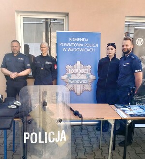 Pośrodku policyjny baner, po prwej i po lewej stronie umundurowany policjant i uczennice klas mundurowanych.