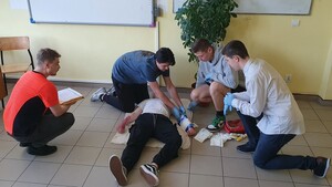 Trzech chłopaków opatruje leżącego na podłodze pozoranta. Po lewej stronie ratownik medyczny. osoby są w pomieszczeniu.