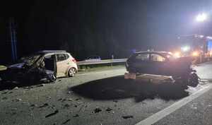 ogólny widok wypadku drogowego na którym są dwa samochody posiadające zniszczenie przedniej części. W okół rozsypane plastikowe elementy, z tyłu samochód straży pożarnej oświetlający miejsce wypadku