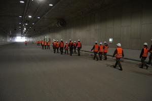 Grupa młodzieży szkolnej ubrana w mora wojskowe a na nich kamizelki odblaskowe barwy pomarańczowej używająca białych kasków budowlanych. Podążają w kierunku wyjścia z tunelu drogowego.