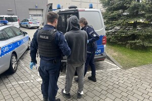 osoba zatrzymana ma założone kajdanki zespolone prowadzona jest do radiowozu przez umundurowanych policjantów