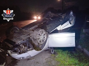 rozbity biały volkswagen leżacy na dachu częsciowo na drodze a częściowo na ogrodzeniu prywatnej posesji