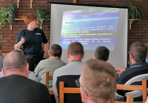 Na zdjęciu widać osoby siedzące na krzesłach są tyłem  Po lewej stronie stoi umundurowana policjantka i patrzy w stronę ekranu na którym jest wyświetlana prezen