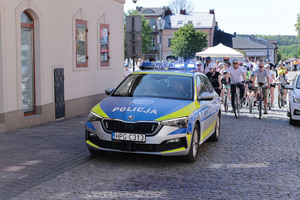 Oznakowany radiowóz ruchu drogowego pilotuje peleton rowerzystów. W tle zabudowania i olkuski Rynek.