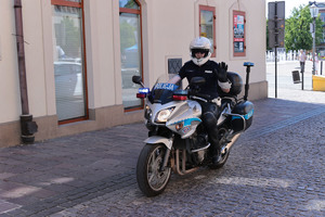 Policjant ruchu drogowego na motocyklu. W tle fragment olkuskiego Rynku i zabudowania