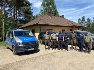 Policjanci i funkcjonariusze strazy leśnej przy pojazdach słuzbowych. w tle budynek oraz krajobraz leśny