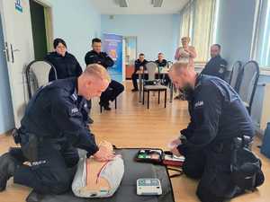 Policjanci ćwiczą pierwszą pomoc  na fantomie przy użyciu defibrylatora. W tle inni funkcjonariusze.