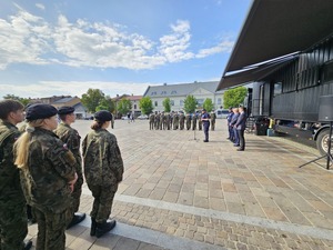 Uczniowei w mundurach w szyku stoją na wprost pojazdu escapetruck. Przed pojazdem przemawia komendant. za nim przedstawieciele władzi i pojazd. W tle zabudowania i olkuski Rynek