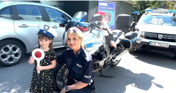 policjantka wraz z dziewczynką trzymającą w ręku tarczę do zatrzymywania pojazdów, chłopiec siedzący na policyjnym