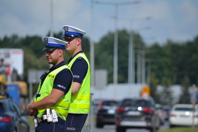 Dwóch policjantów ruchu drogowego stoi przy drodze i nadzoruje ruch. W tle wzmożony ruch na drodze