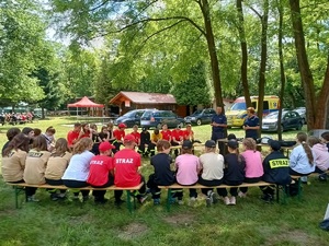 Grupa osb siedzących tyłem na ławkach, przed nimi umundurowana policjnatka i policjnat. W tle namiot,drewniany buynek, grupa osób siedzącch na ławkach i drzewa.