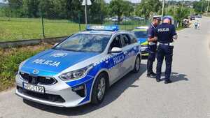 Po lewej stronie dwa radiowozy stojące jeden za drugim obok nich dwaj policjanci. W tle zaparkowane pojazdy i ludzie.