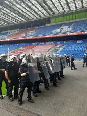 umundurowani policjanci wyposażeni w tarcze ochronne stojący przy płycie boisko skierowani w kierunku jednej z trybun stadionu