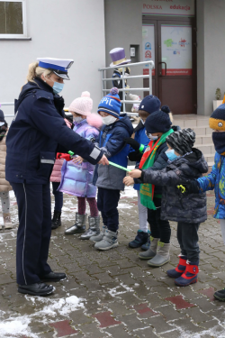 nr 1 – policjantka ruchu drogowego wręcza dzieciom elementy odblaskowe , w tle wejście do budynku szkoły. Dzieci stoją w grupie ubrane w maseczki.