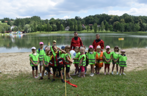 Dzieci w kamielkach odblaskowych oraz osoboy proawdzące pokaz na tle zalewu wodnego