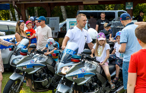 Policjant stoi przy policyjnych motocyklach, na których siedzą dzieci. Obok uczestnicy pikniku