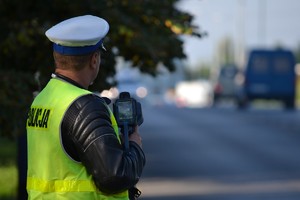 Policjant ruchu drogowego stoi przy krawędzi jezdni - dokonuje pomiaru prędkości ręcznym miernikiem.