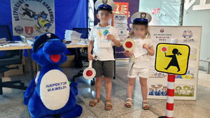 dwoje dzieci ubranych w policyjne czapki, obok policyjna maskotka, za nimi policyjne stoiusko