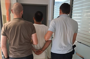 dwóch nieumundurowanych policjantów prowadzi zatrzymnaego mężczyznę z złożonymi kajdankami na rękach trzymanych z tyłu przez korytarz komisariatu jpg