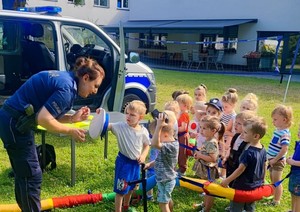 Miejski Żłobek w Oświęcimiu policjantka prezentuje dzieciom czapkę w tle radiowóz
