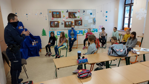 Grupa dzieci siedzących na krzesłach, oddalonych od siebie, wśród nich opiekunka grupy. Po lewej stronir stoi umundurowany policjant w maseczce założonej na nos i usta. Osoby znajdują się w ładnie udekorowanej klasie.