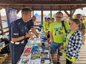 Stanowisko profilaktyczne policji. Dwóch policjantów, przy stolikach. Na przeciwko czestnicy imprezy, dzieci z rodzinami. Na stolikach ulotki, plakaty broszury, kolorowe malowanki, .