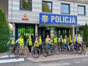 Policjant wraz z dziećmi trzymającymi rowery stoi przezd wejściem do komendy policji