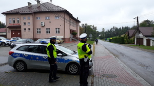 Policjant z miernikiem prędkości kontroluje prędkość jadących pojazdów na drodze. Obok stoi drugi policjant i radiowóz