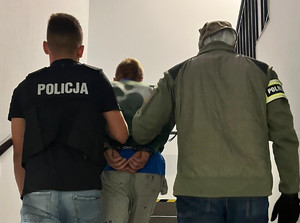 policjanci prowadzą zatrzymanego z kajdankami na ręce trzymane z tyłu po klatce schodowej w komendzie