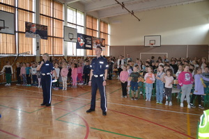 Policjantka i policjant pokazują licznej grupie uczniów na sali gimnastycznej  jak należy przechodzić przez jezdnię