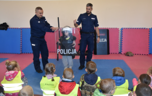Policjanci oraz przedszkolaki podczas prelekcji. jeden z uczniów trzyma policyjną tarc oraz ma na sobie kamizelkęzę
